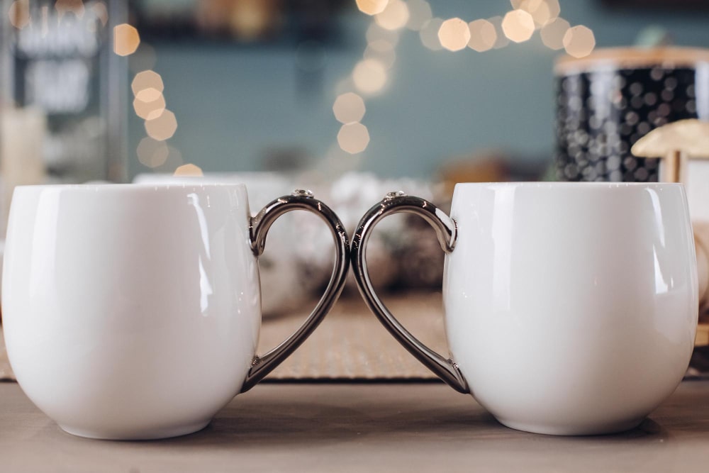 Duo de mugs blancs aux anses formant un cœur, posés sur une table en bois avec des lumières bokeh en arrière-plan