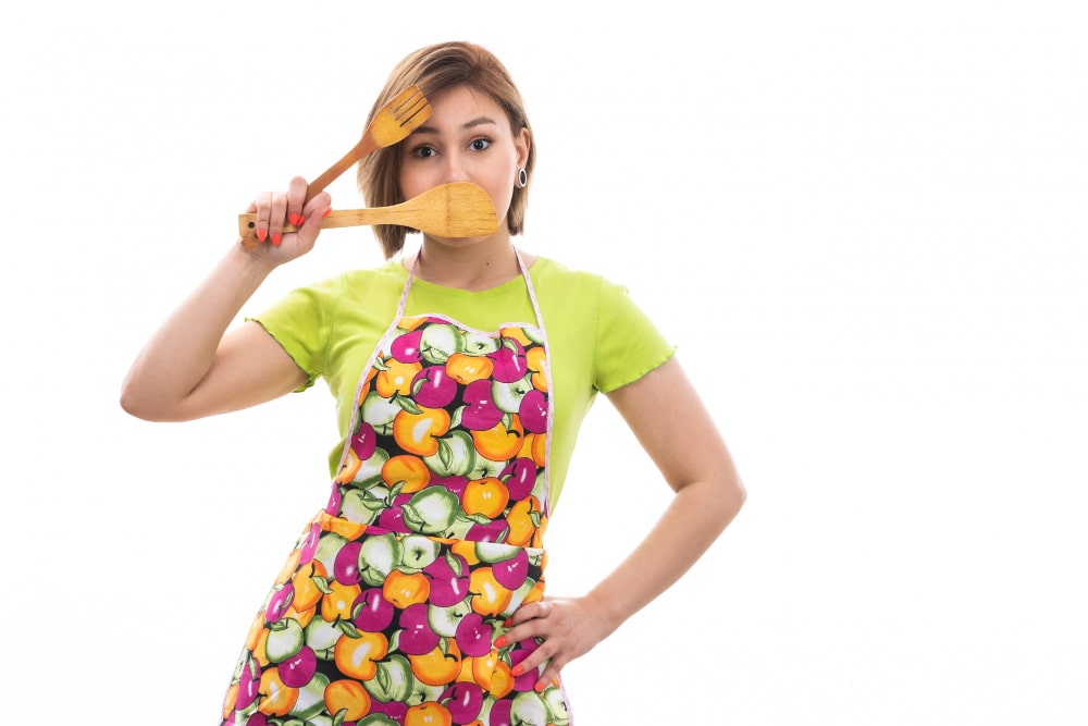 Jeune femme portant un tablier humoristique aux motifs fruits colorés et jouant avec des ustensiles en bois
