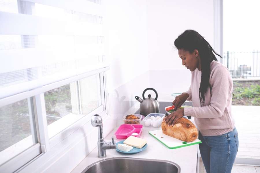 Une femme prépare un repas dans une petite cuisine blanche lumineuse avec plan de travail compact et fenêtre donnant sur l'extérieur.