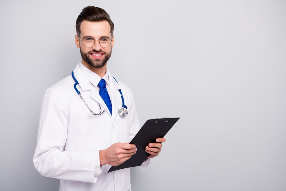 Portrait d'un jeune docteur souriant en blouse blanche tenant un presse-papiers sur fond gris.