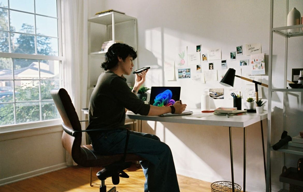 Un jeune créatif utilise une commande vocale sur son smartphone tout en travaillant sur son ordinateur portable, illustrant l'usage quotidien des applications intelligence artificielle.