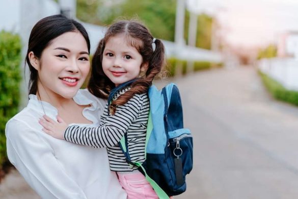 Une femme sourit en portant dans ses bras une petite fille équipée d'un sac à dos bleu.