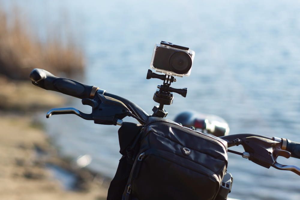 Une caméra insta360 en moto et à vélo installée sur un guidon à l'aide d'un support de fixation, face à un plan d'eau ensoleillé.