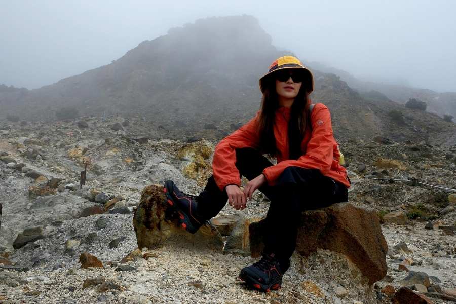 Une randonneuse assise sur un rocher, portant une veste orange et un bob, prend une pause contemplative sur un sentier de montagne rocheux et brumeux, illustrant l'esprit du slow tourisme.