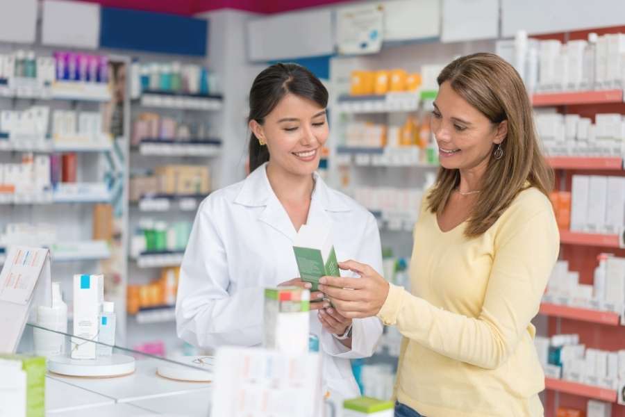 Une pharmacienne souriante conseille une cliente qui tient une boîte de médicaments dans une officine moderne.